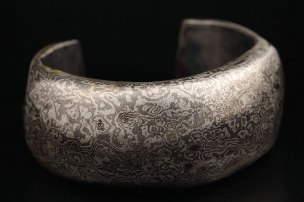 Wide Mokume Gane cuff, its silver surface patterned like wood grain, on a dark background